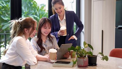 Three women enjoying coffee while looking at a tablet in a modern office.