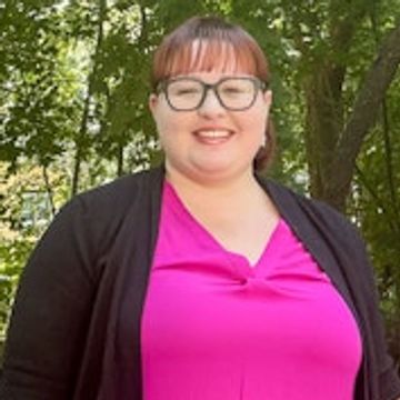 Smiling woman with glasses in a bright pink top and black cardigan outdoors.