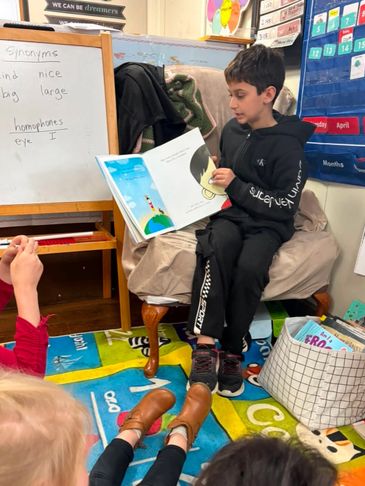 A boy reads a book aloud to classmates in a colorful classroom.