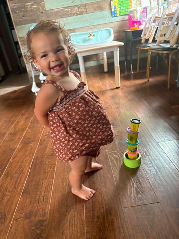 A happy toddler stands barefoot on a wooden floor next to colorful stacking cups.