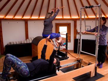 Two women exercising on Pilates machines inside a wooden room with an instructor.
