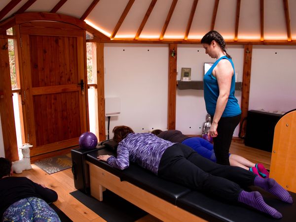 A Pilates instructor assists a woman lying on a reformer machine.