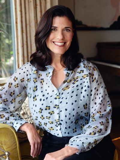 Woman smiling while seated indoors in a patterned blouse.