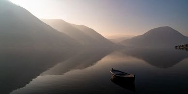 A solitary boat floats on calm lake waters surrounded by misty mountains at dawn.