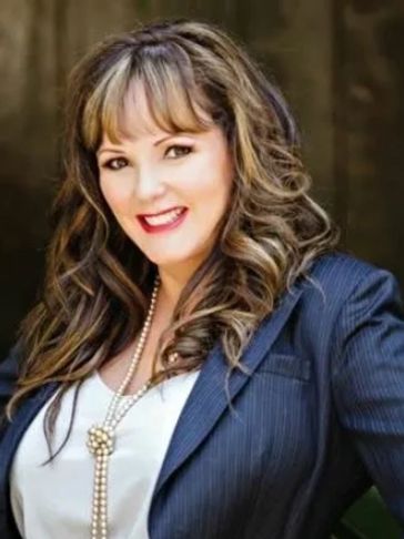 A confident woman with curled hair wearing a blazer and pearl necklace.