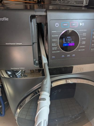 A washing machine with an open detergent drawer and a vacuum cleaner hose inserted.