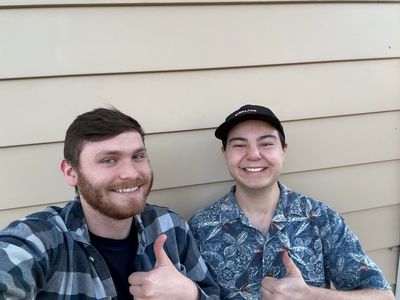 Two smiling men giving thumbs up, one in a plaid shirt and the other in a floral shirt and cap.