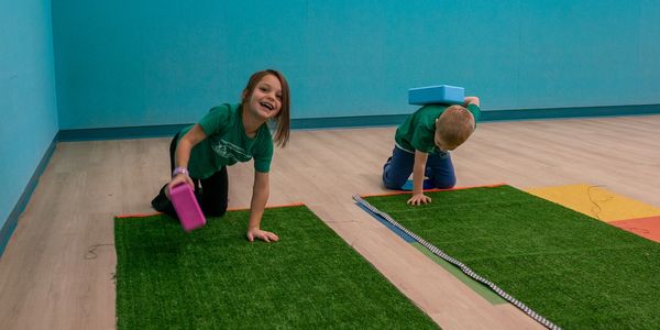 Two children play balancing games on green mats in a room with blue walls.