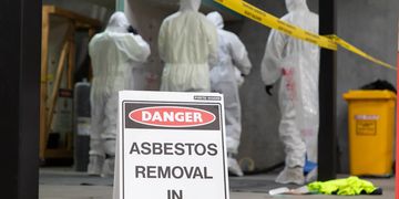 Workers in protective gear removing asbestos behind a danger sign.