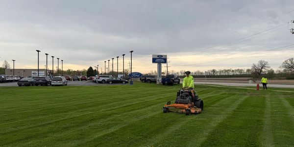 Worker mowing grass at a Ford dealership on a cloudy day.