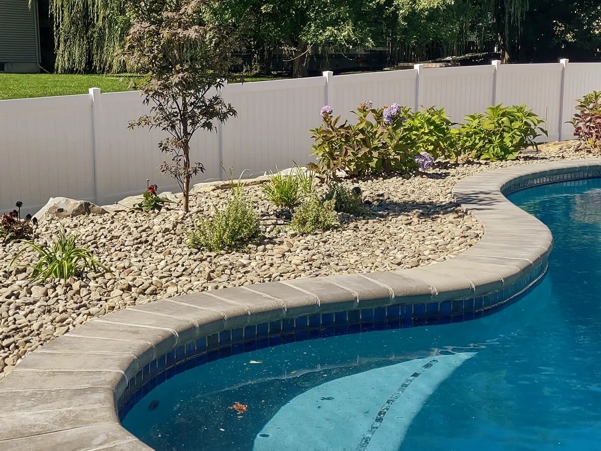 White vinyl privacy fence along a landscaped backyard pool with stone patio edge and planting beds.