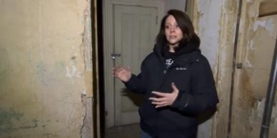 Woman in a dark jacket explaining inside a worn, old building.