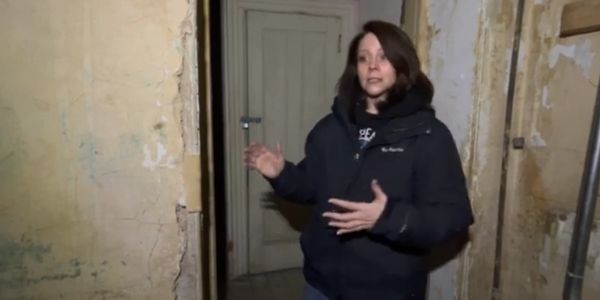 Woman in a dark jacket explaining inside a worn, old building.