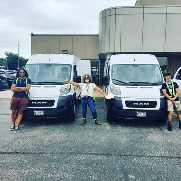 Three people standing in front of two white RAM vans in a parking lot.