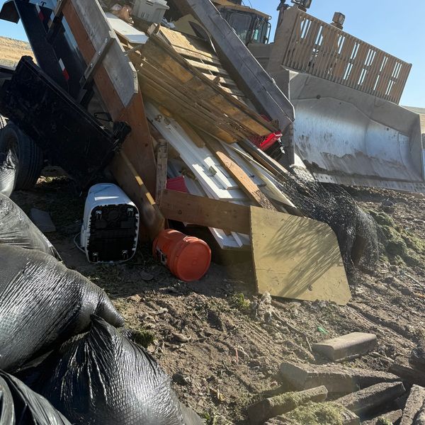 Construction debris and trash bags piled on a dirt ground near heavy equipment.