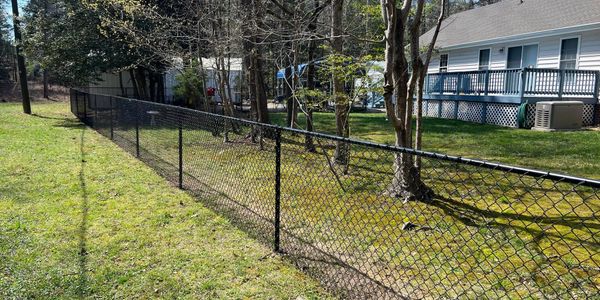 Black chain-link fence dividing two grassy yards with trees and houses.
