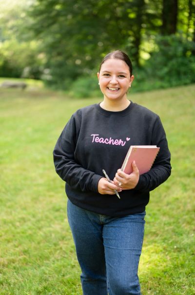 Smiling woman wearing a teacher sweatshirt in a park.