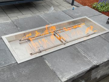 Modern outdoor fire pit with flames on a stone patio near lounge chairs.