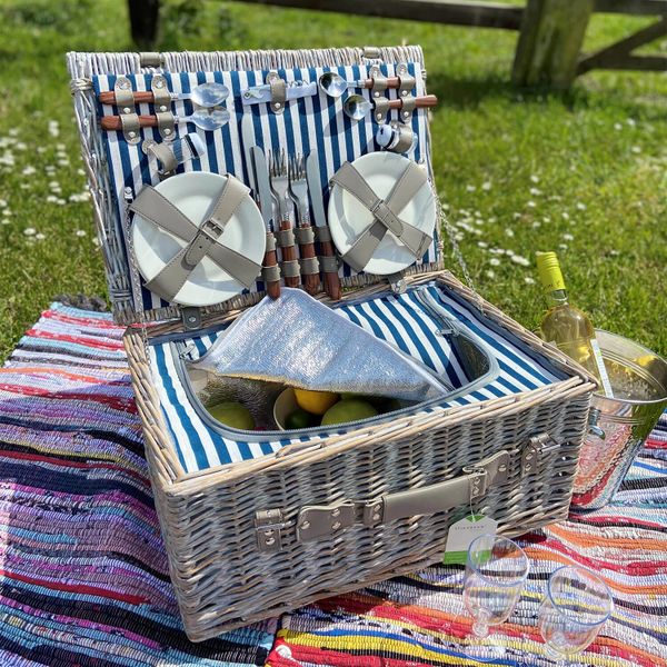 A picnic basket with plates, cutlery, fruits, and wine on a colorful blanket outdoors.