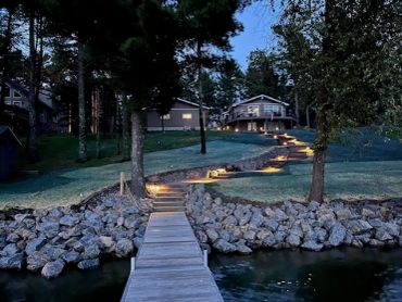 A wooden dock leading to a lit pathway and houses at dusk.