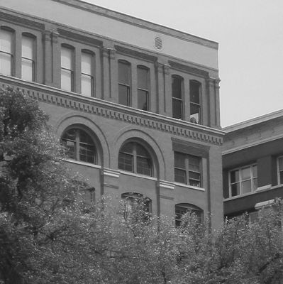 Black and white image of an old brick building with arched windows and trees in front.