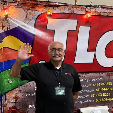 Man with mustache waving in front of colorful wall mural.