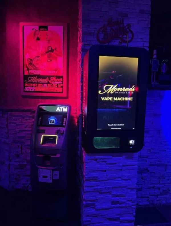 ATM and vape machine glowing under neon lights in a dimly lit setting.