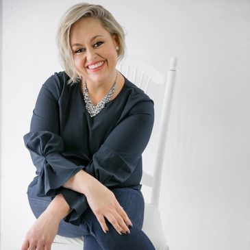 Smiling woman seated on a white chair, dressed in a dark blouse and statement necklace.