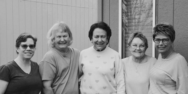 Five smiling women standing side by side outdoors in black and white.