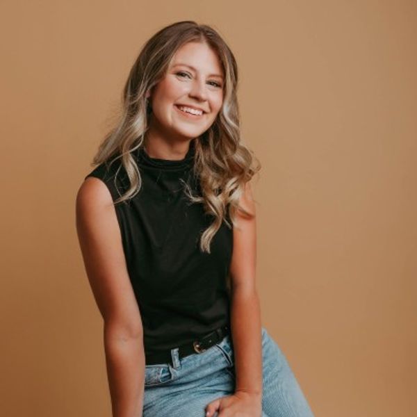 Smiling woman in black sleeveless top and jeans against a brown background.