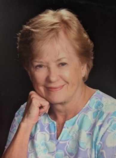 Smiling elderly woman with light hair in a floral blouse, resting her chin on her hand.