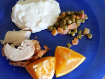 A blue plate with mashed potatoes, peas and carrots, turkey, and orange slices.