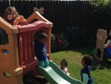 Children playing on a small plastic slide in a sunny backyard.