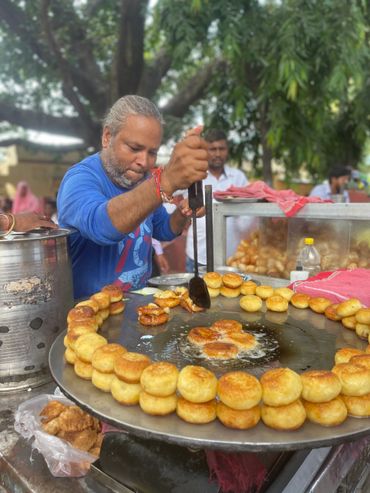 A street vendor frying and arranging golden brown snacks on a large circular griddle.