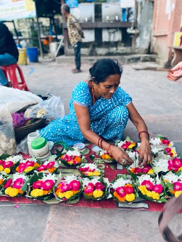 A woman arranges colorful flower offerings on the ground at a market.
