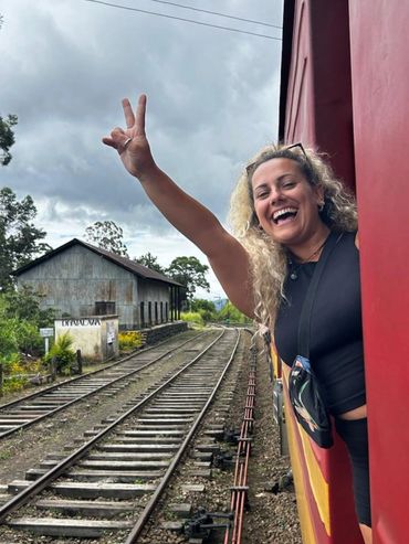 A joyful woman leans out of a train window, waving with a peace sign.