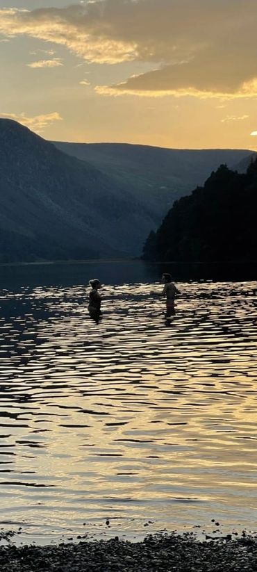 Two people stand in a calm lake at sunset surrounded by mountains.