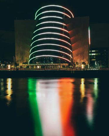 Modern building illuminated with red, white, and green lights reflecting on water at night.