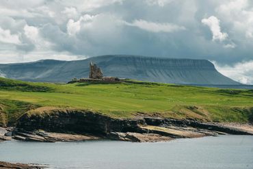 Ruins on a grassy cliff by the sea with a mountain backdrop under cloudy skies.