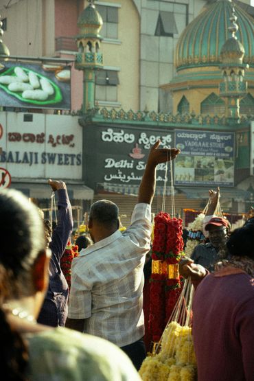 A bustling market scene with a man holding flower garlands in front of traditional Indian shops.