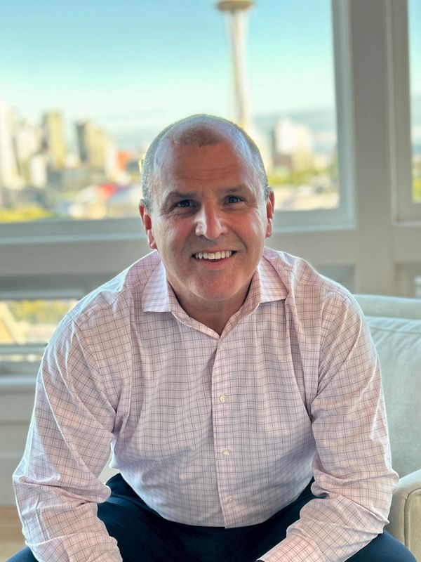 Smiling man in a checkered shirt sitting indoors with a cityscape background.