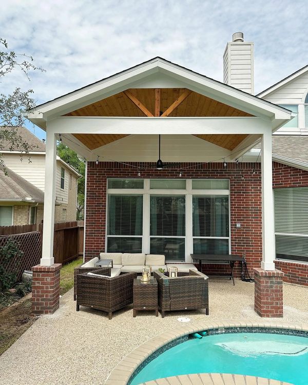 Covered patio with wicker furniture next to a swimming pool at a brick house.