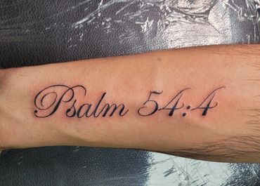 Tattoo of 'Psalm 54:4' in elegant script on forearm.