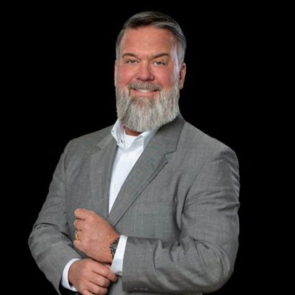 Confident man with gray beard in a gray suit smiling against a black background.