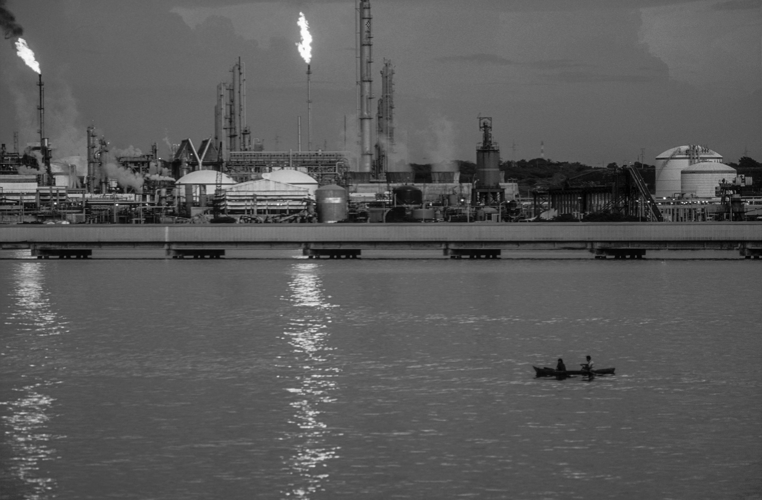 Industrial refinery with flames and a boat on water in foreground.