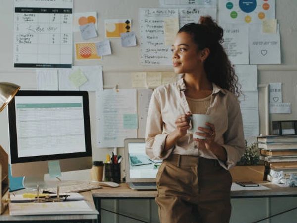 Woman holding a cup, standing thoughtfully in her workspace with a computer and notes on the wall.
