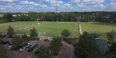Aerial view of a large soccer field with cars parked and people walking around.