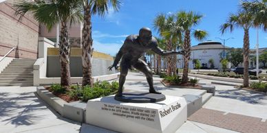 Bronze statue of a surfer in a sunny outdoor plaza with palm trees.