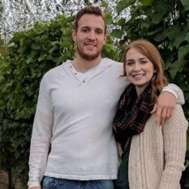 A smiling couple posing outdoors with greenery behind them.
