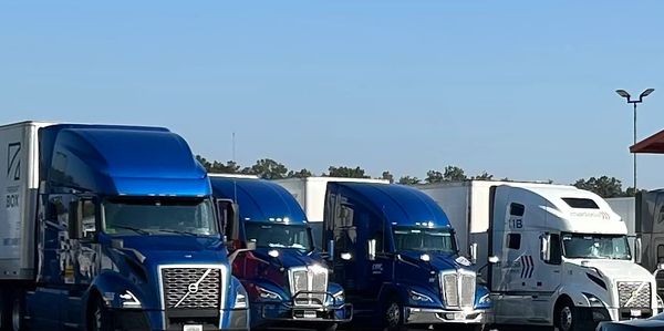 Four large semi-trucks parked side by side under clear blue sky.